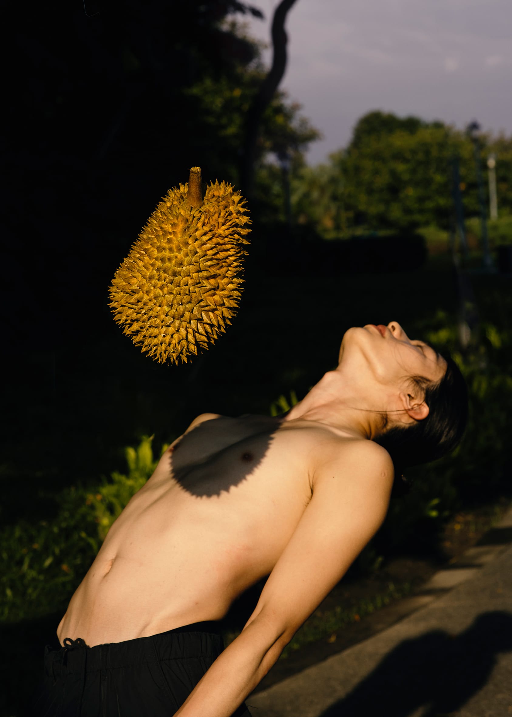 A shirtless person, head tipped backward, eyes closed is cast in sunlight. A fruit appears to be falling on their chest.