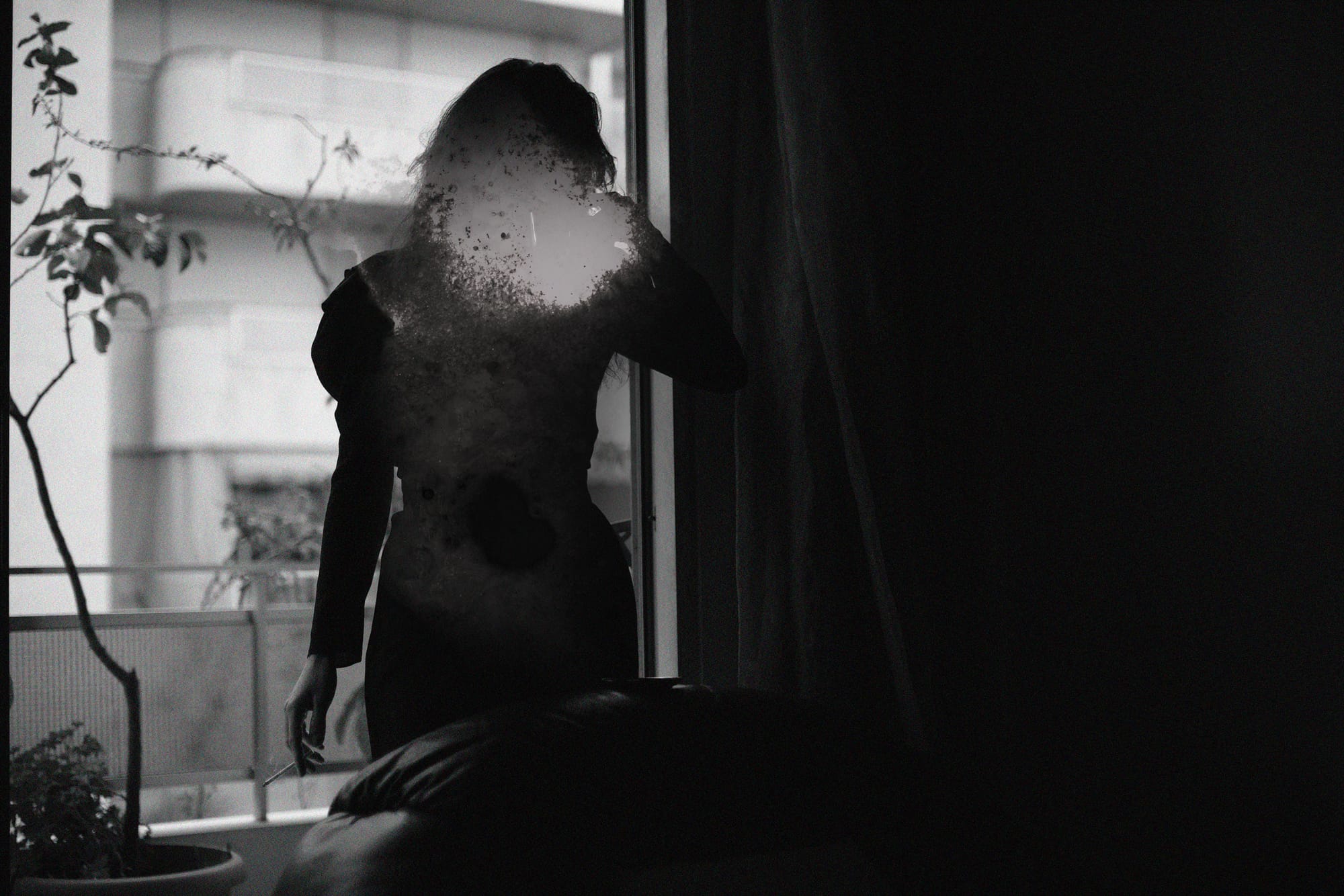 A black and white photo of a woman at a window. Her face is fully obscured.