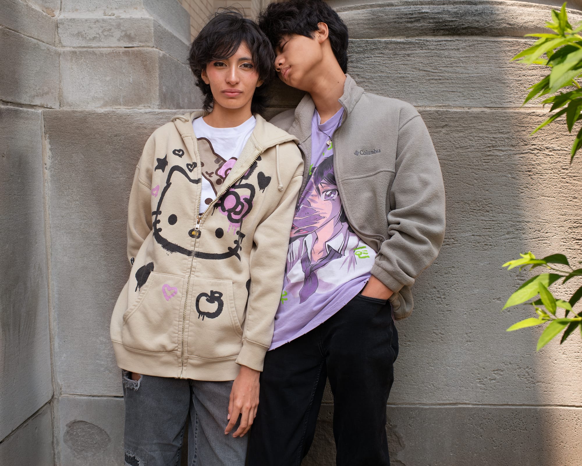 Two individuals lean against a wall; one leans in toward the other. They are wearing T-shirts featuring Hello Kitty and a manga character.
