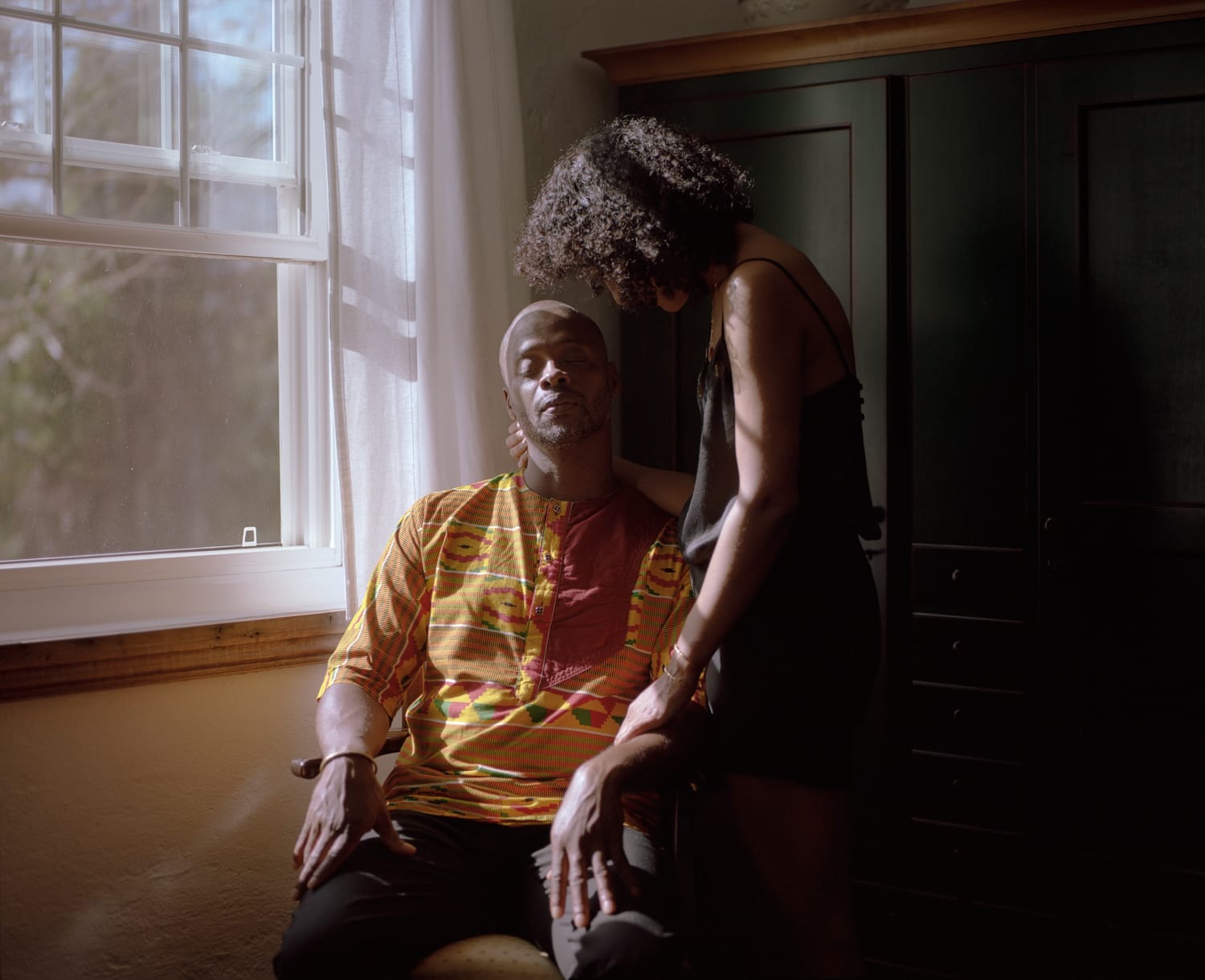 A man sits in a chair with his eyes closed. A woman stands next to him, one hand on his arm.