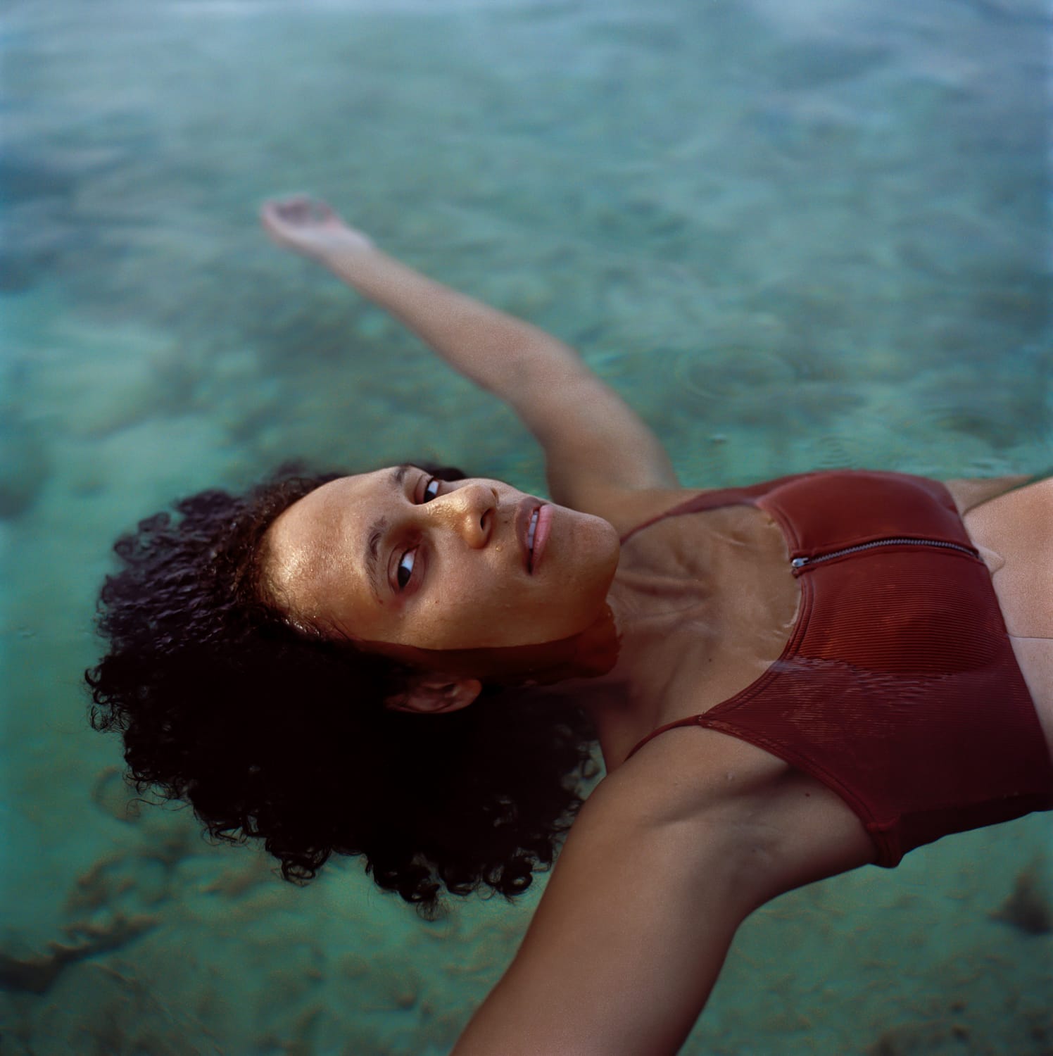 A woman in a red bikini top floats on the water, her eyes looking into the camera.