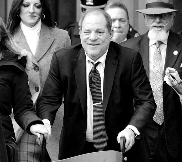 Harvey Weinstein wearing a suit and using a walker, surrounded by other people walks towards the camera