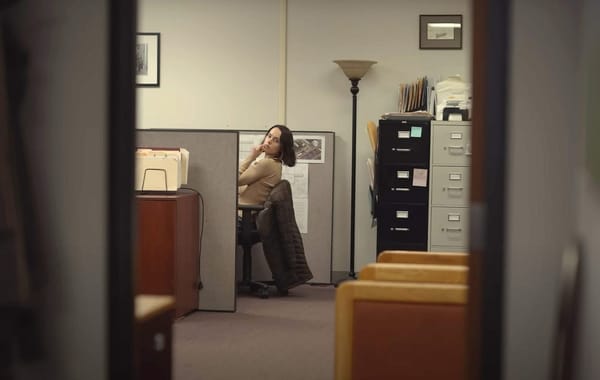 A woman with short, dark hair leans out of her cube in a beige office. 