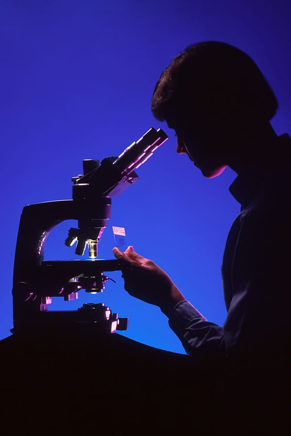 A woman is silhouetted against a blue background as she peers into a microscope.