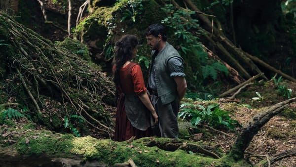 In still taken from the movie Hamnet, Agnes and Will stand in a forest, close together, as though they are about to kiss.