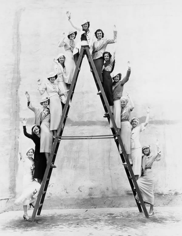 Black and white side-on shot of an A-frame ladder. Women, who look to be from the 1940s. are standing on every other rung, smiling and waving. 