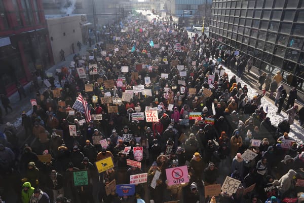 Photo of a large crowd in a built-up area. They carry signs including "ICE Out!", "GTFO", "Do You Hear The People Sing?" and "No Kings, No ICE". 