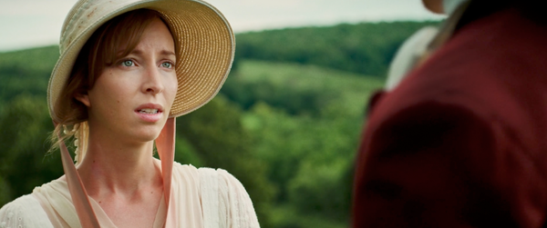 Still from Jane Austen's Period Drama. Close up of Miss Estrogenia, wearing a wicker bonnet. In the foreground is a shoulder clad in red velvet (p. In the background are rolling green hills.