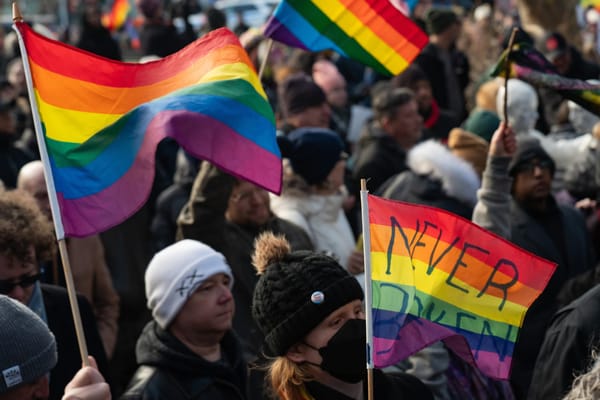 Photo of a stoney-faced crowd waving rainbow Pride flags. One flag has 'NEVER BROKEN' written on it.