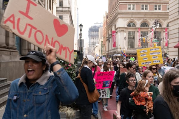 Photo of a women's march. In the foreground, a woman waves a sign: 'I heart autonomy'. In the background, someone carries another: 'If only children were protected like the Epstein Files'.